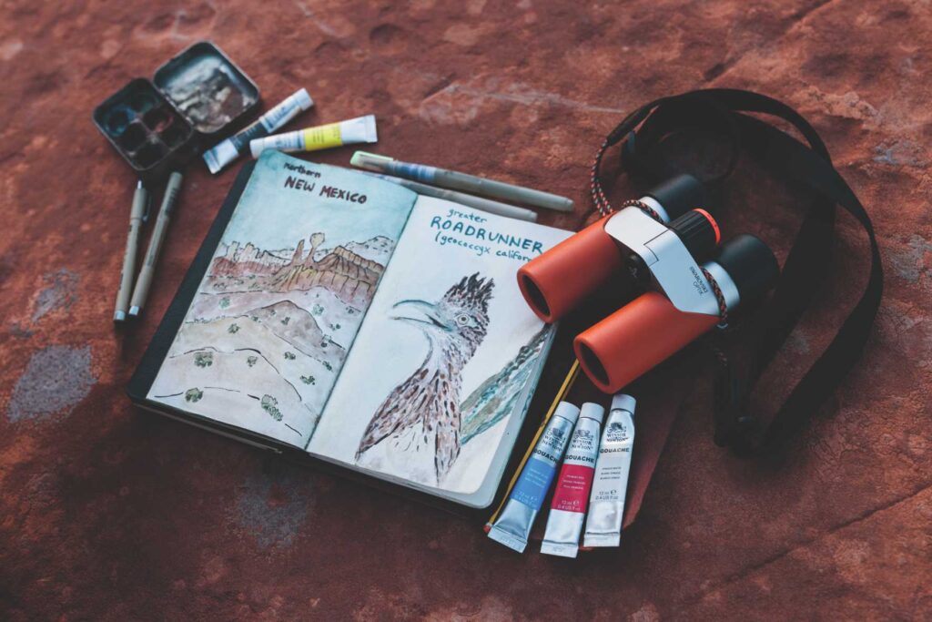 Swarovski CL Fernglas in Desert Orange neben einem Reisetagebuch und Malutensilien.