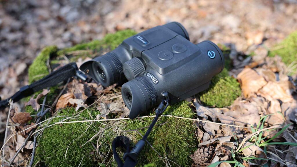 Steiner eDiscovery 10x42 Fernglas liegt auf moosbewachsenem Baumstamm im Wald zwischen Laub und Zweigen.