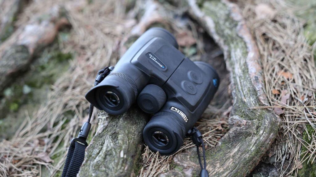 Steiner eDiscovery 10x42 Fernglas liegt zwischen Baumwurzeln und Nadeln im Wald, Detailaufnahme von oben.